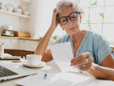 Beltéri felvétel egy elgondolkodó, zavart, ősz hajú, szemüveges idős hölgyről, aki a kezével a fejét támasztva a fizetési csekket nézi, és a magas lakbér miatt bosszús, miközben a konyhaasztalnál ül a laptopja előtt