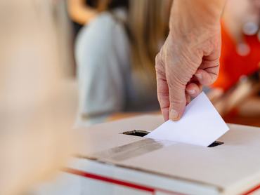 Close-up of hand placing ballot into voting box at election. Ballot and voting box symbolize democracy and elections. Hand placing ballot in election voting box, casting a vote at election