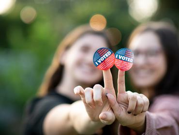 Portrait of two women  holding out their hands together with the I voted stickers attached to their fingers for the viewer to see. Their fingers form the v for victory and peace sign.