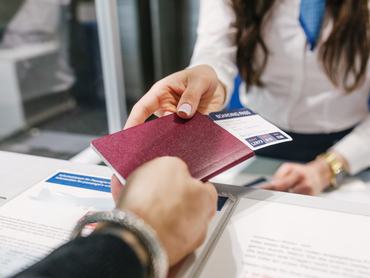 Közelkép egy útlevélről és beszállókártyáról, amelyeket egy női repülőtéri alkalmazottnak adnak át, miközben egy férfi bejelentkezik a járatára.