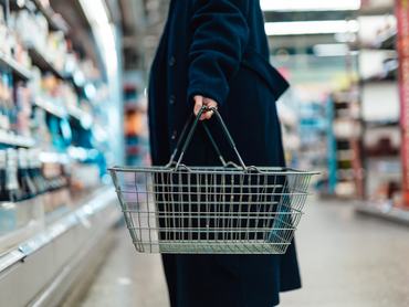 Közelkép egy bevásárlókosarat tartó fiatal nőről, aki a szupermarket zöldség-gyümölcs részlegénél áll.