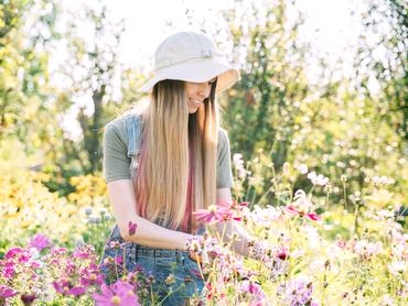 Egy nő ollóval nyírja a növényeket. Egy kalapos, színes kesztyűs fiatal nő gondozza a virágokat a kertben.