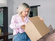 Rossz csomag. Egy szomorú, csalódott középkorú hölgy kicsomagolja a dobozt, elégedetlen a rossz kézbesítési szolgáltatással, miközben otthon a nappaliban ül a kanapén. Szállítási hiba